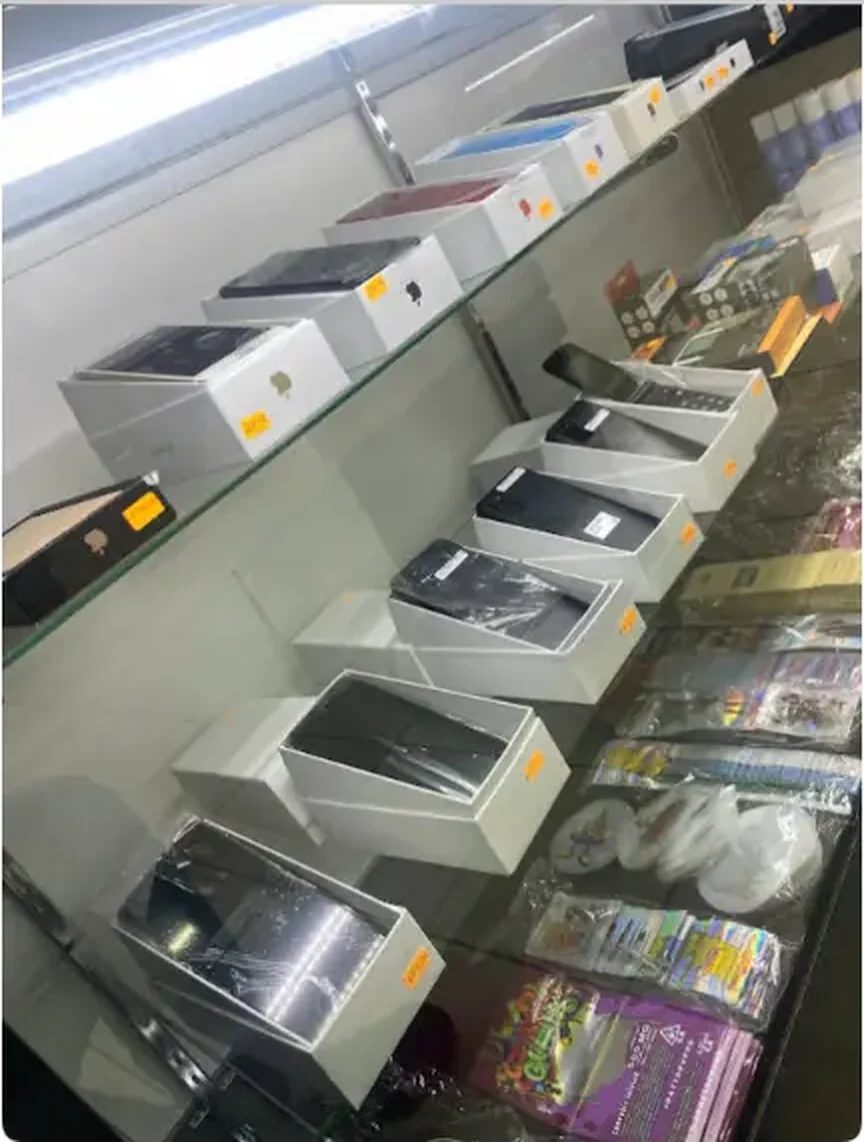 Mega Tobacco Vape & Wireless glass shelf of boxed smartphones and tablets beside packaged accessories